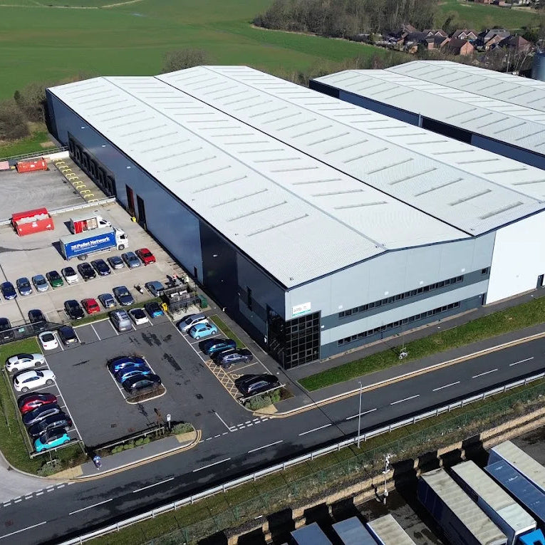 Aerial view of a large industrial warehouse complex with a white roof, gray walls, a parking lot with cars, and a road with trucks in the foreground.